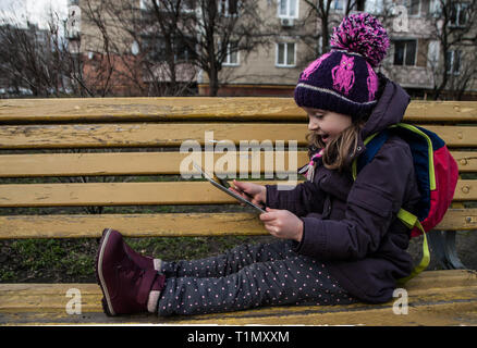 Beautiful little girl in purple jacket and knit hat sitting on yellow shabby bench and holding digital tablet. Happy smiling child has fun with gadget Stock Photo