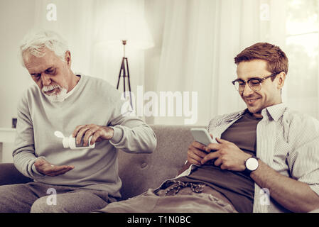 Indifferent grandchild looking at phone meanwhile his granddad taking pills. Stock Photo