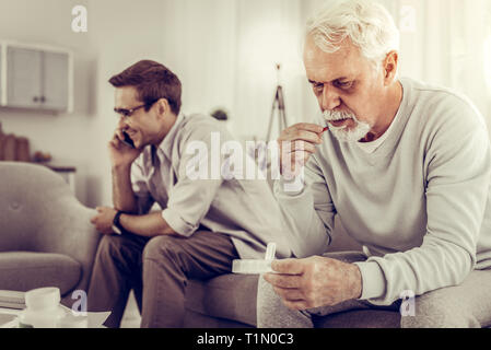 Old man taking pills while son indifferently having a phone call. Stock Photo