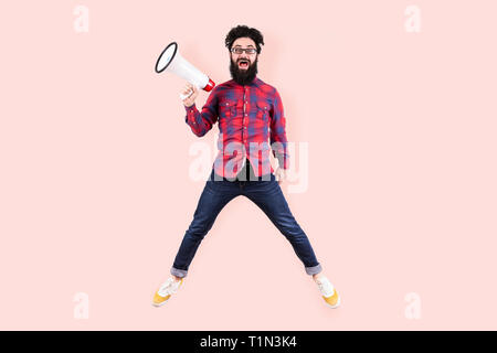 full length studio portrait of bearded crazy hipster man in glasses, jumping and shouting in megaphone and looking at the viewer, photo on pink backgr Stock Photo