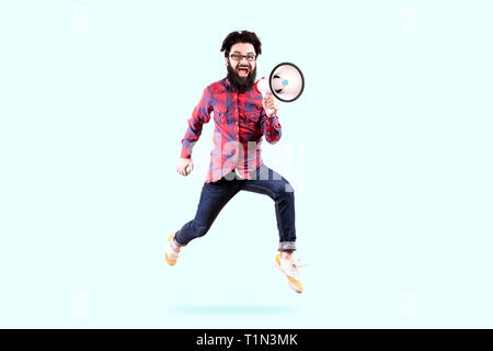 Bearded hipster man in glasses, jumping and shouting in megaphone, photo on blue background Stock Photo