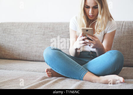 Attractive young woman relaxing on a sofa at home and using a touch screen smart phone to socialize and network from home Stock Photo