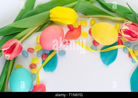 Colorful Easter wreath made of yellow tulips, colorful eggs and candies on white background. Flat lay. Stock Photo