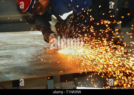 A worker in gloves and overalls cuts a metal sheet with an angle-grinding machine. Sparks fly from the disk. Industrial background with space for copy Stock Photo