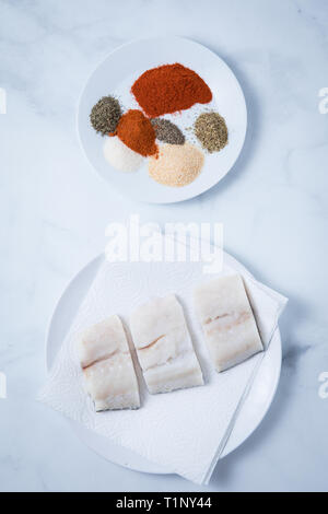 Raw cod fish filets on napkins and a round white plate with spices on a white marble background. Uncooked pieces of cod. Stock Photo