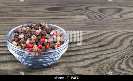 Peppercorns mix. Black, white, green and red pepper corns. Glass bowl on wooden background. Piper nigrum. Colorful mixture of dried seasoning close-up. Stock Photo