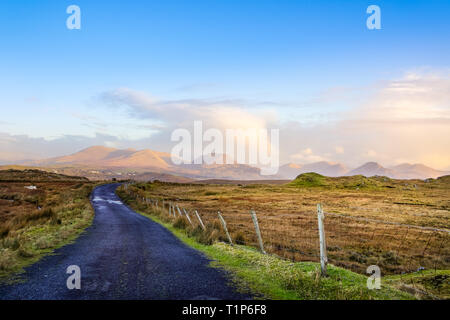 Empty country road between meadows leading to mountains in far distance, blue sky with puffy clouds in golden hour Stock Photo