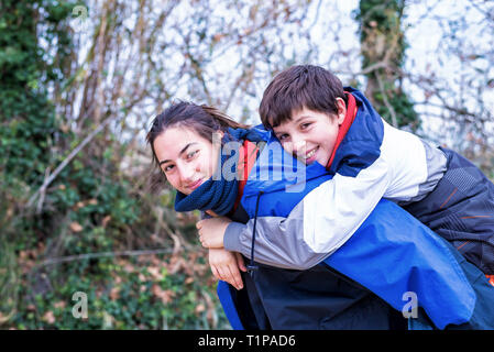 Young boy piggyback on his sister looking at camera and smiling in nature Stock Photo