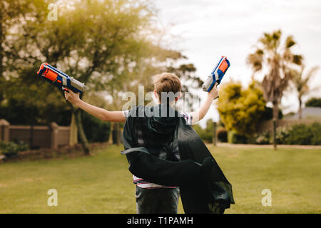 Rear view of boy wearing a black cape and running with two toy guns in hand. Young boy playing dressed up in cape. Stock Photo