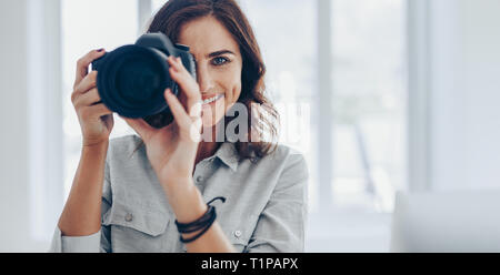 Happy young woman taking pictures with her DSLR camera indoors. Professional photographer taking photos in her studio. Stock Photo