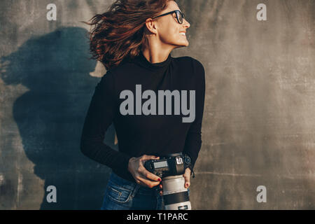 Beautiful female photographer with a professional camera against muslin backdrop. Cheerful young woman photographer with digital camera. Stock Photo