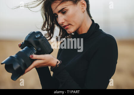 Photographer checking pictures on dslr camera. Young woman wearing casuals standing outdoors with professional camera. Stock Photo
