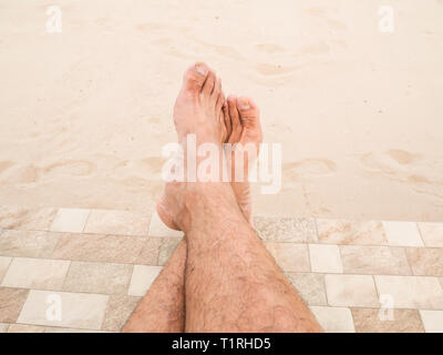 Man is relaxing barefoot at the beach. Closeup legs of Asia man's skin and men hairy legs on sand background. Guy's legs on the sand. Stock Photo