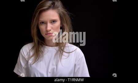 Portrait of young caucasian long-haired girl in shirt watching aggressively into camera on black background. Stock Photo