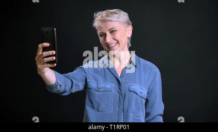 Closeup portrait of adult cheerful caucasian female with short blonde hair making selfies on the phone and smiling in front of the camera Stock Photo