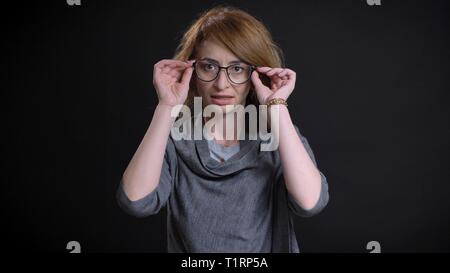 Closeup portrait of middle-aged extravagant redhead female looking straight at camera fixing her glasses with background isolated on black Stock Photo