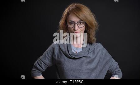 Closeup portrait of middle-aged extravagant redhead female in glasses having hands on the hips and posing while looking straight at camera Stock Photo