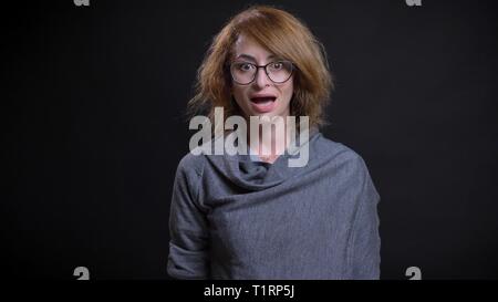 Closeup portrait of middle-aged extravagant redhead female in glasses getting surprised and excited while looking straight at camera Stock Photo