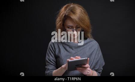 Closeup portrait of middle-aged extravagant redhead female in glasses texting on the tablet in front of the camera with background isolated on black Stock Photo