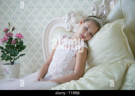 Cute tender girl teenager lies on the bed. Princess in a white dress with a crown. Stock Photo