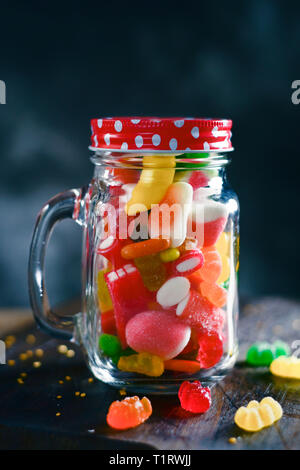 a glass mason jar full of candies with different flavors and colors, on a rustic wooden table, against a dark background Stock Photo