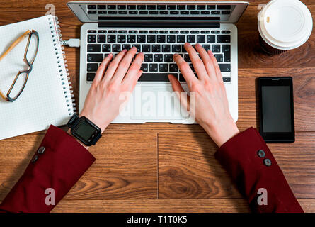 Business person working at office desk, typing and using laptop, top view. Smart watch on hand and smart phone on the table. Coffee cup, notepad Stock Photo