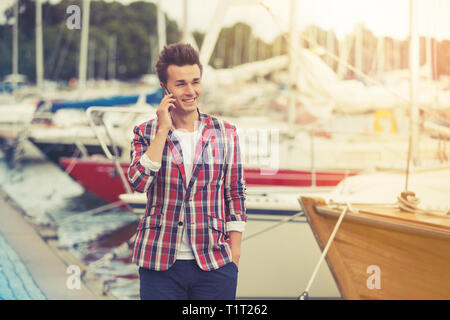 Portrait of handsome smiling stylish man in plaid blazer walking along sea bay. Talking on the phone. Sailboats in harbour. Street style concept. Stock Photo