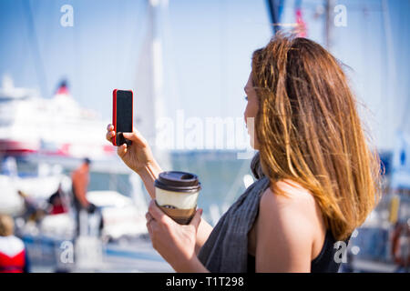 Beautiful woman taking  photo with smart phone. Picture of harbour with sailboats, seaside. Sunny summer bay. Taking selfie and drinking coffee. Stock Photo