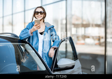 Lifestyle portrait of a young beautiful woman in blue coat using phone near the modern car outdoors Stock Photo