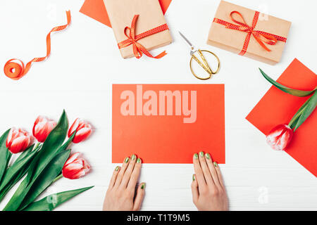 Top view of female's hands holding blank red paper card near gift boxes and bouquet of tulip flowers on white wooden table. Stock Photo