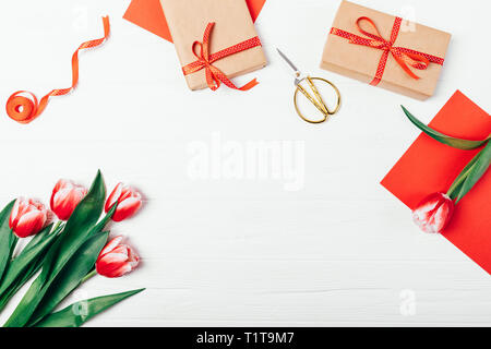 Spring decorative frame of beautiful tulip flowers, gift boxes and red cards on white wooden table, top view. Stylish flat lay composition with empty  Stock Photo