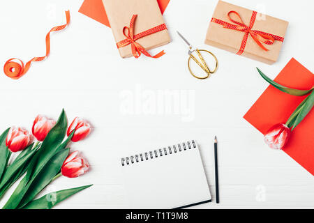 Spring decorative frame of beautiful red tulip flowers and gift boxes on white wooden table, top view. Stylish flat lay composition with blank notepad Stock Photo