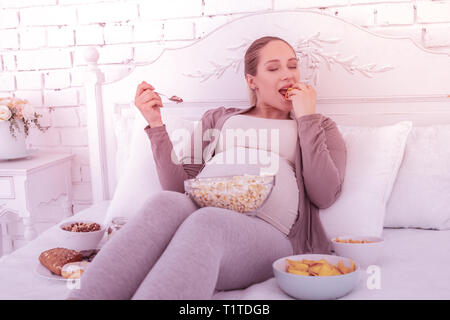 Fair-haired good-looking pregnant woman in comfortable clothes eating with pleasure Stock Photo
