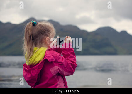 Child taking a travel photo on a mobile phone of a loch and landscape in Scotland Stock Photo