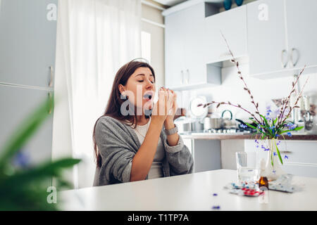 Spring allergy. Young woman sneezing because of flowers surrounded with pills on kitchen at home. Seasonal allergy concept. Stock Photo