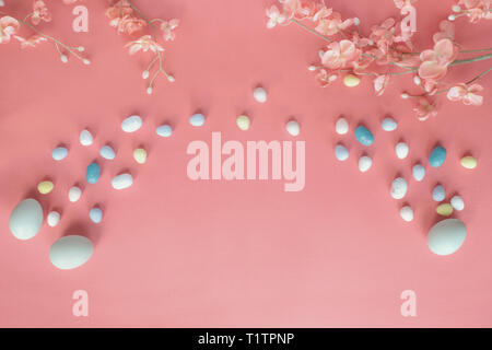 Pastel Easter eggs, malt candy covered chocolate eggs, and flower blossoms over a coral colored background with copy space. Image shot from top view. Stock Photo