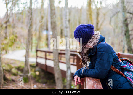 Side view portrait of beautiful girl with backpack standing near old footbridge. She is having break during hike in coniferous forest Stock Photo