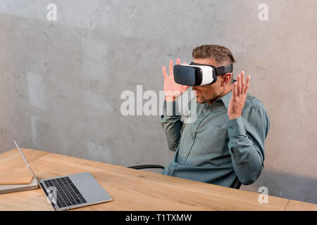 shocked businessman wearing virtual reality headset while sitting near laptop in office Stock Photo