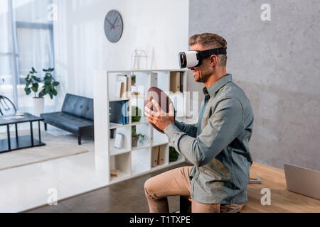 cheerful businessman wearing virtual reality headset while holding american football Stock Photo