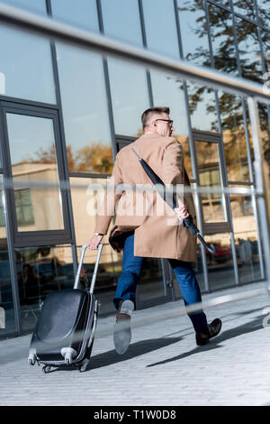 businessman in beige coat running with suitcase and umbrella on street Stock Photo