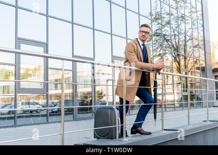 businessman in glasses and beige coat standing with umbrella near suitcase Stock Photo