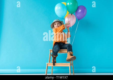 cute kid in jeans and orange shirt sitting on stairs and pointing with finger at balloons Stock Photo