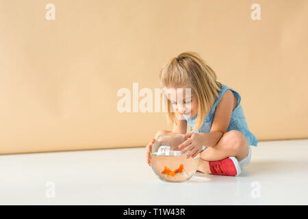 adorable kid sitting with crossed legs and looking at fishbowl on beige background Stock Photo