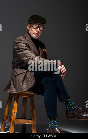 stylish mature businessman in cap and glasses looking at camera on grey Stock Photo