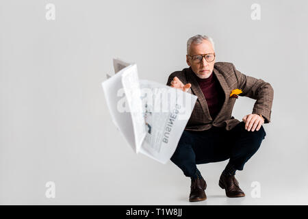 selective focus of mature man throwing in air business newspaper while sitting on grey Stock Photo