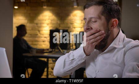 Closeup shoot of adult caucasian businessman typing on the laptop yawning and being exhausted indoors in the office Stock Photo