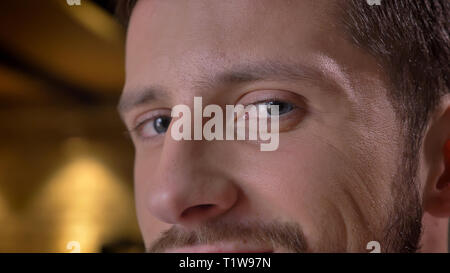 Closeup shoot of adult caucasian bearded male face with eyes looking at camera with smiling facial expression indoors with interior on the background Stock Photo