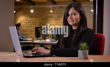 Portrait of middle-aged overweight businesswoman sitting in front of laptop and smiling into camera on office background. Stock Photo