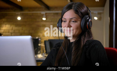 Portrait of middle-aged overweight woman in headphones watching into laptop smilingly on office background. Stock Photo