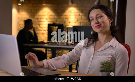 Closeup shoot of young cheerful caucasian businesswoman typing on the laptop smiling and being satisfied indoors on the workplace Stock Photo